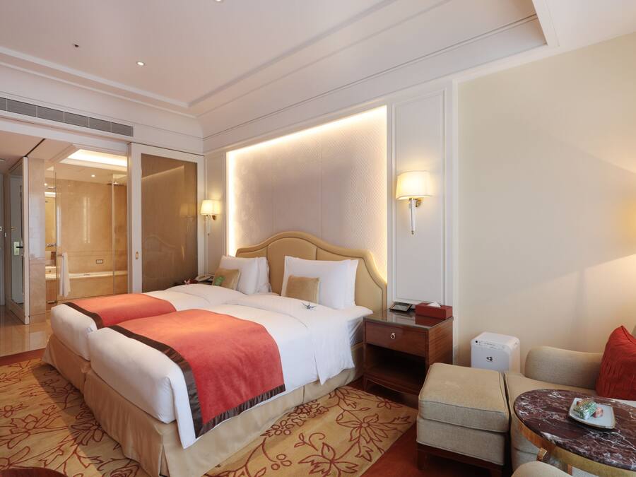 A hotel room with a large bed, bedside tables, a seating area with a chair and a small table, and a bathroom visible through an open door.
