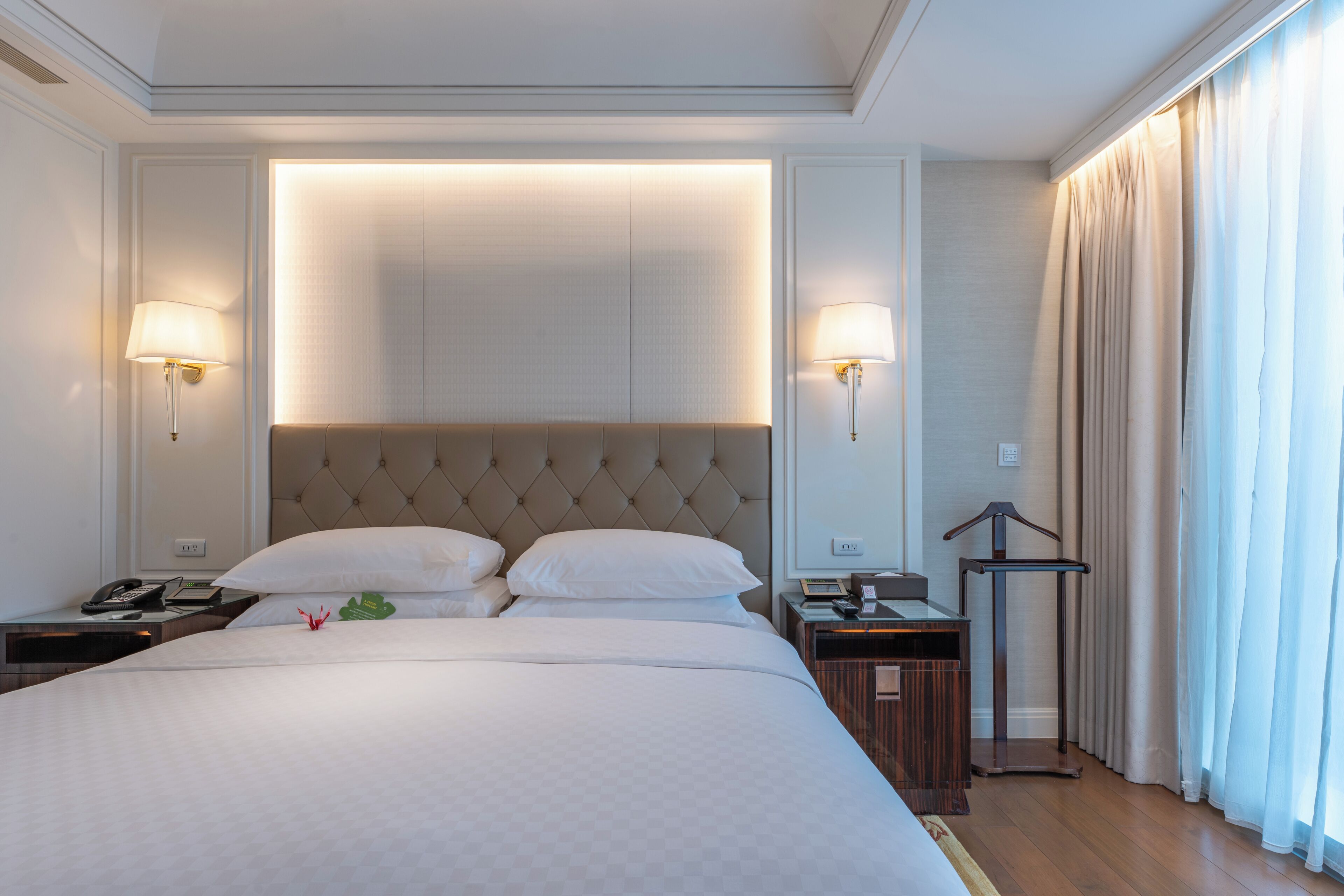 A hotel room with a large bed, two bedside tables, a hanging light fixture, and a window with curtains.