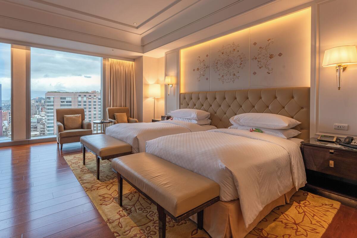 Une chambre d’hôtel avec deux lits, un banc, une chaise, une table de chevet, une lampe et une grande fenêtre donnant sur la ville.