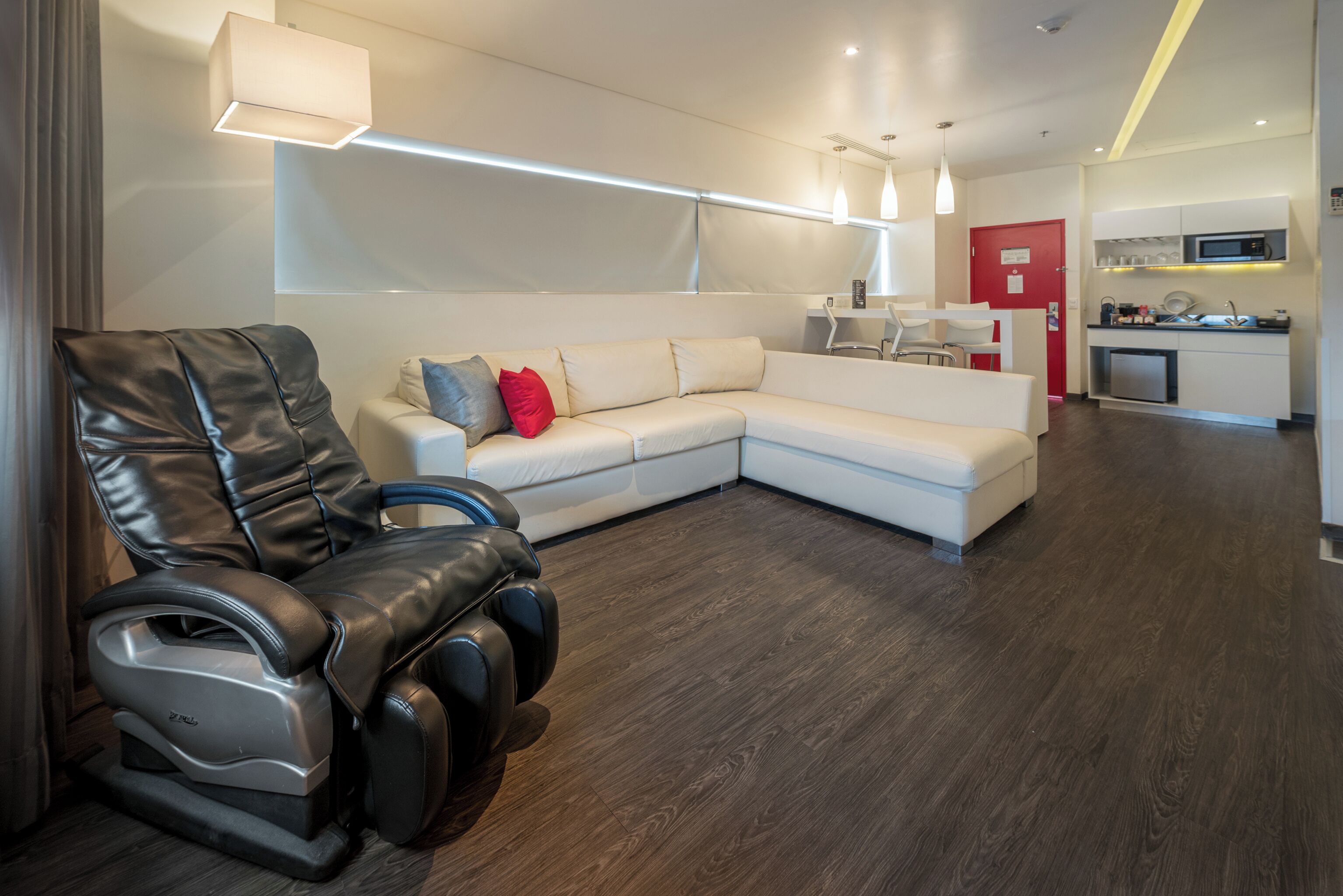 A modern living room with a leather recliner, a white sectional sofa, and a red door leading to a kitchen area.