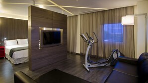 A modern hotel room with a large bed, a TV mounted on a wooden panel, an exercise bike, and a large window with curtains. - Ramada Encore by Wyndham Guadalajara Sur (Tlajomulco de Zúñiga)