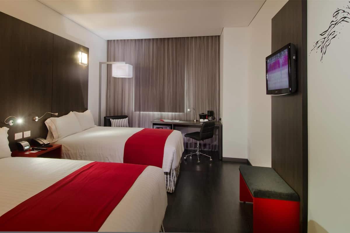 A hotel room with two beds, a desk, a chair, a TV, and a window with curtains.
