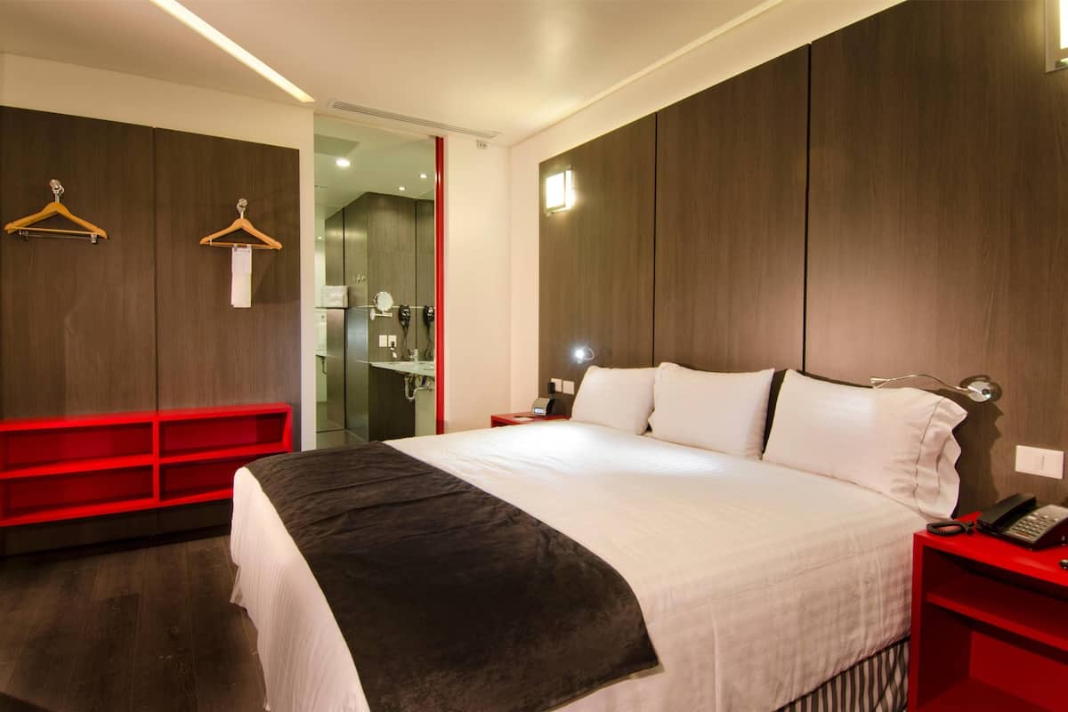 A modern hotel room with a large bed, a red nightstand, a red bench, and wooden paneling on the walls.