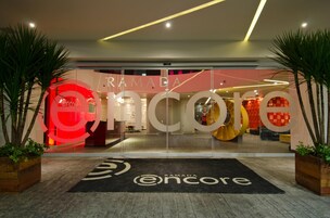 A modern hotel entrance with a glass facade, a large sign reading "RAMADA oncore," and a black doormat with the hotel's logo.