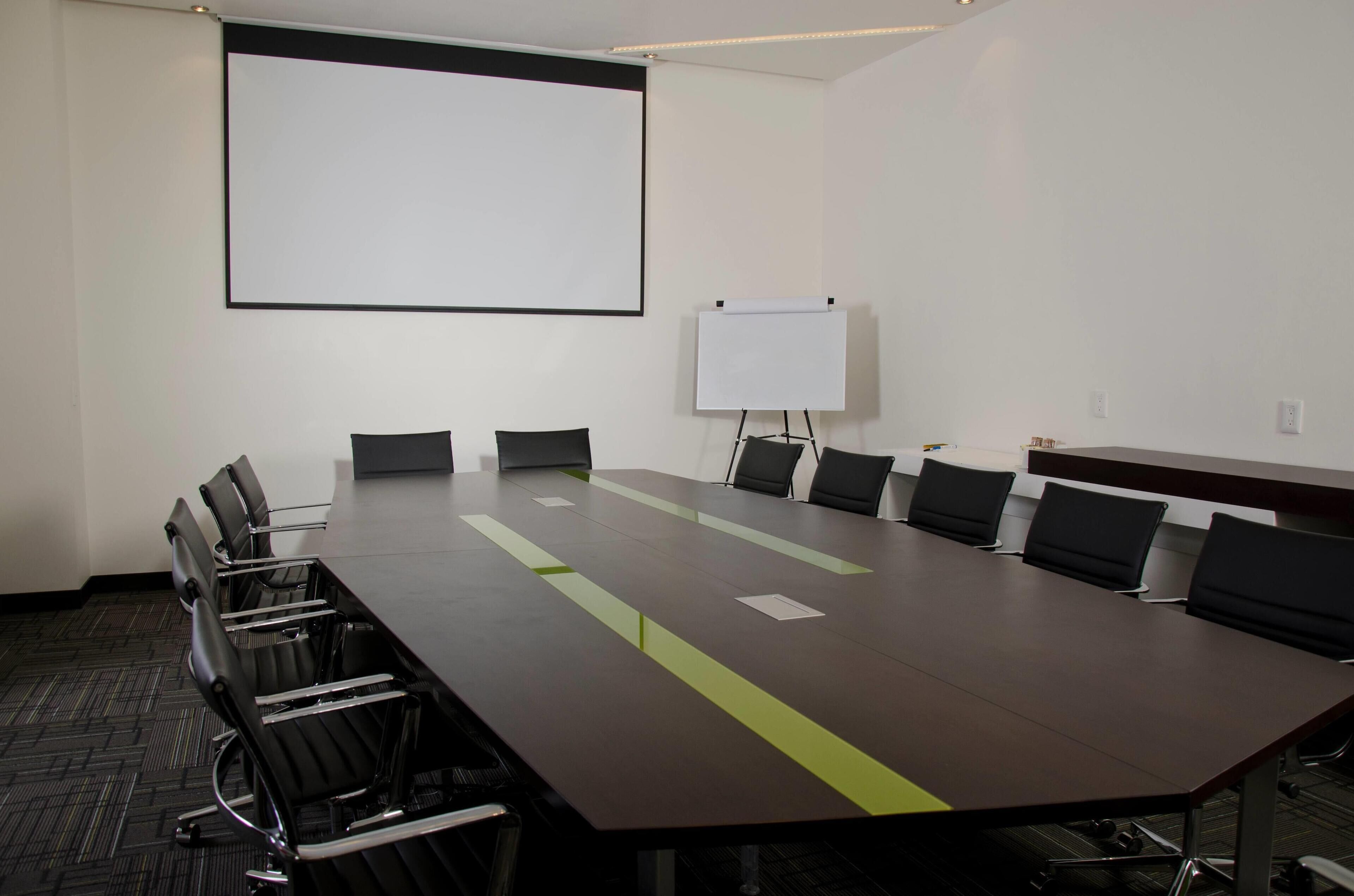 Een vergaderruimte met een lange tafel, zwarte stoelen, een whiteboard en een projectiescherm.