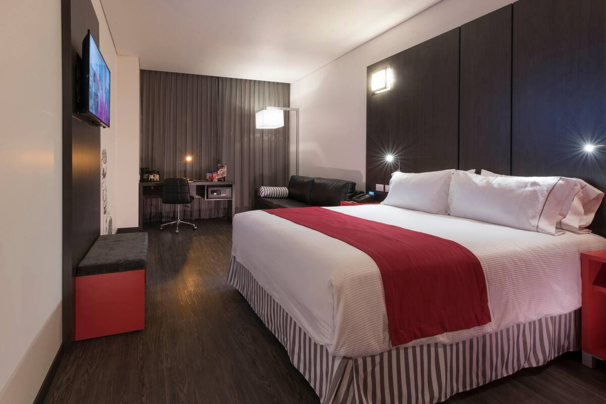 A modern hotel room with a large bed, a desk, a chair, and a TV on the wall.