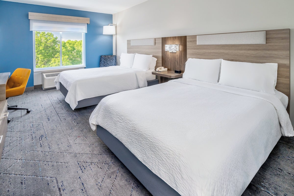 A hotel room with two beds, a desk, a chair, and a window with a view of trees.