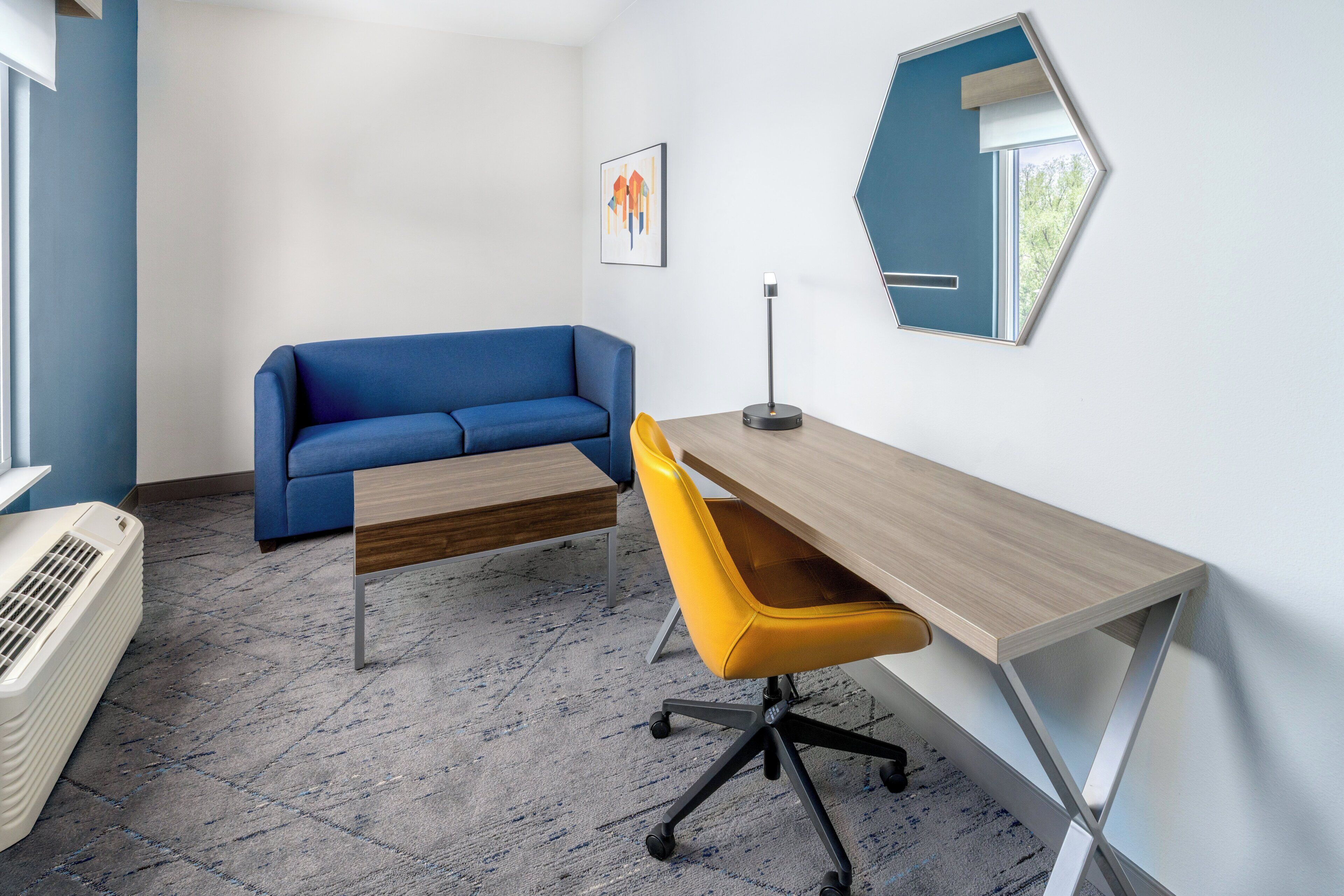 Un espacio de oficina moderno con un sofá azul, un escritorio de madera, una silla amarilla y un espejo hexagonal llamativo.