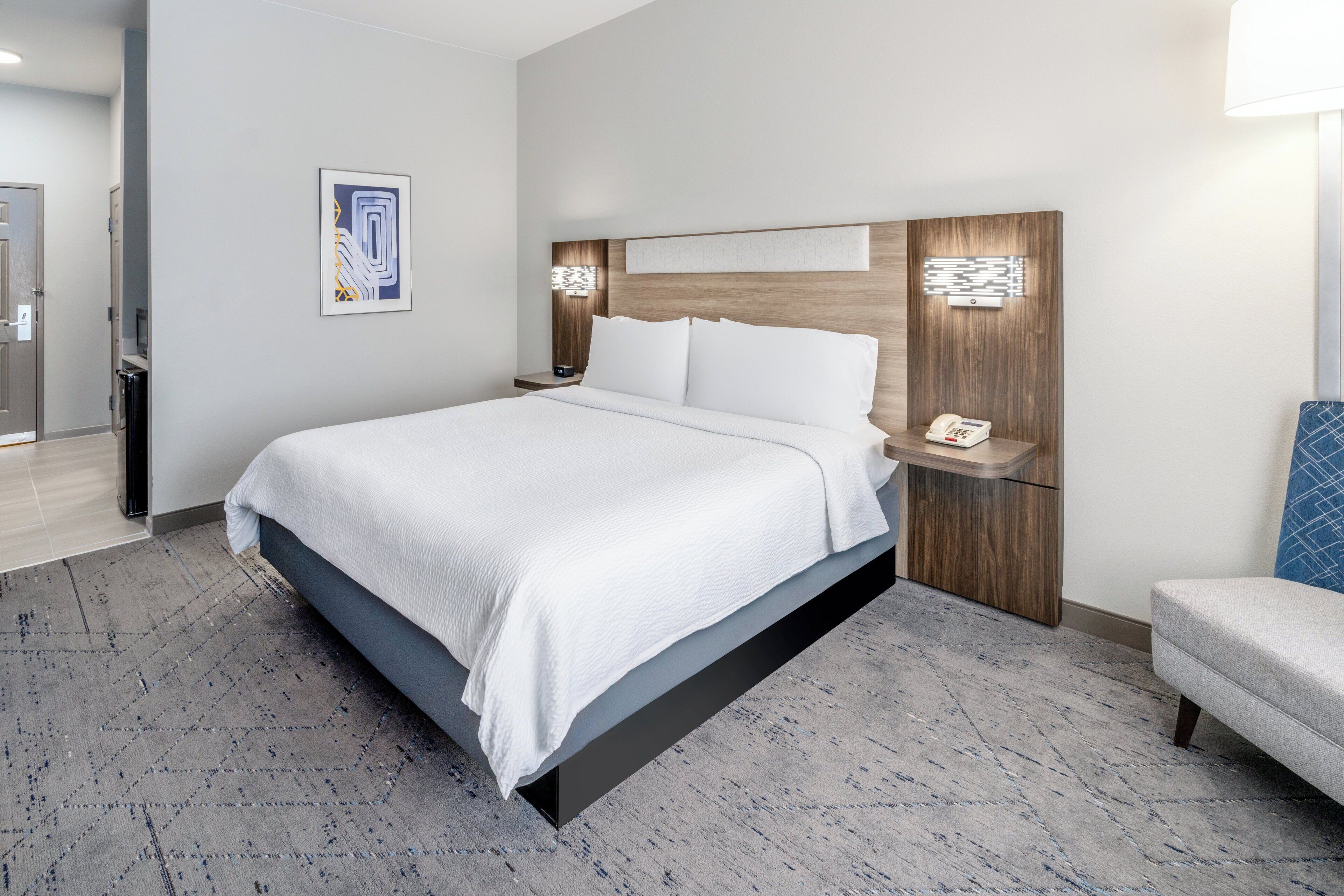 Una habitación de hotel moderna con una cama grande, una mesita de noche con lámpara y un pequeño rincón de descanso.