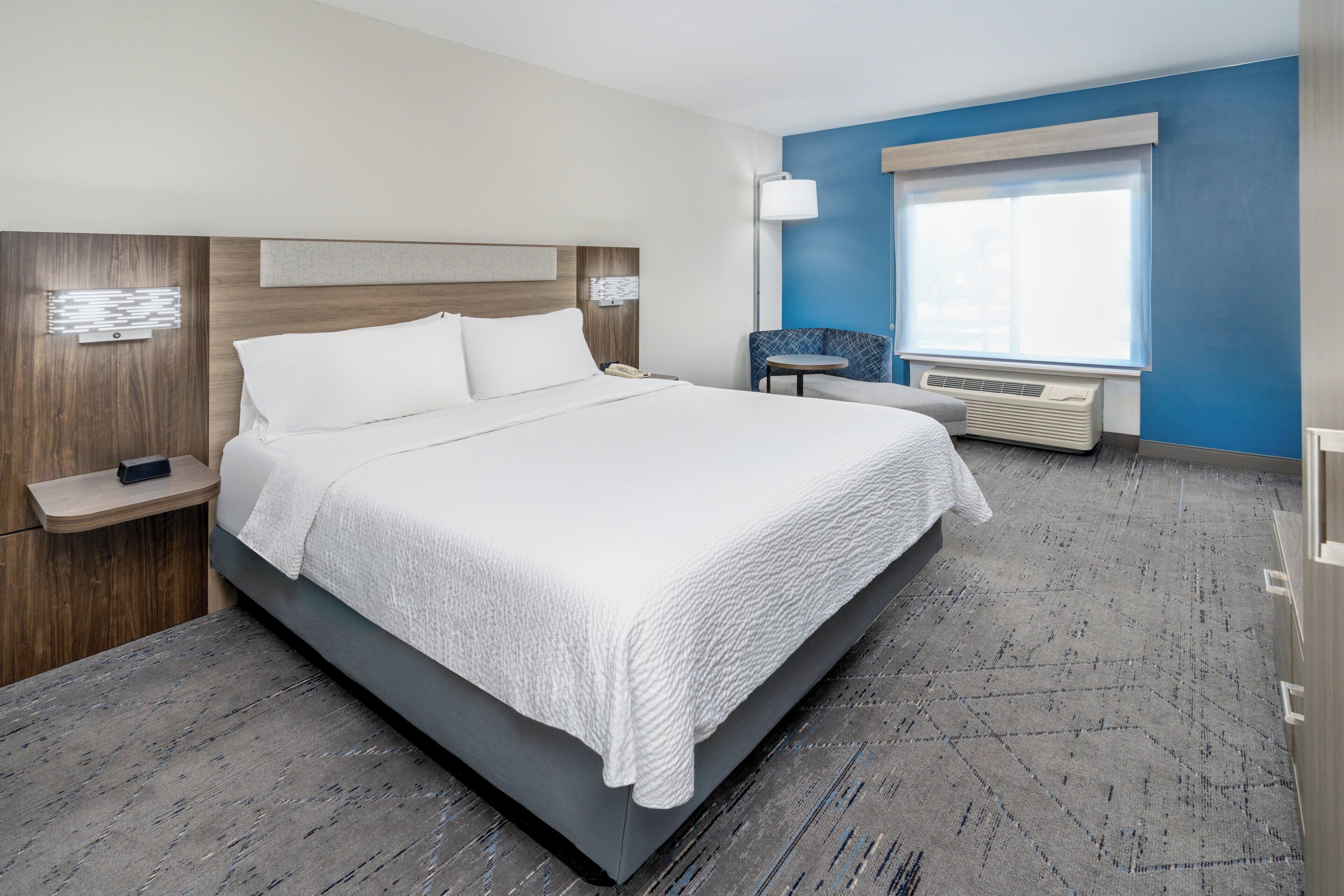 A modern hotel room with a large bed, bedside tables, a chair, a window with blinds, and an air conditioning unit.