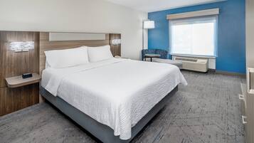 A modern hotel room with a large bed, bedside tables, a chair, a window with blinds, and an air conditioning unit.