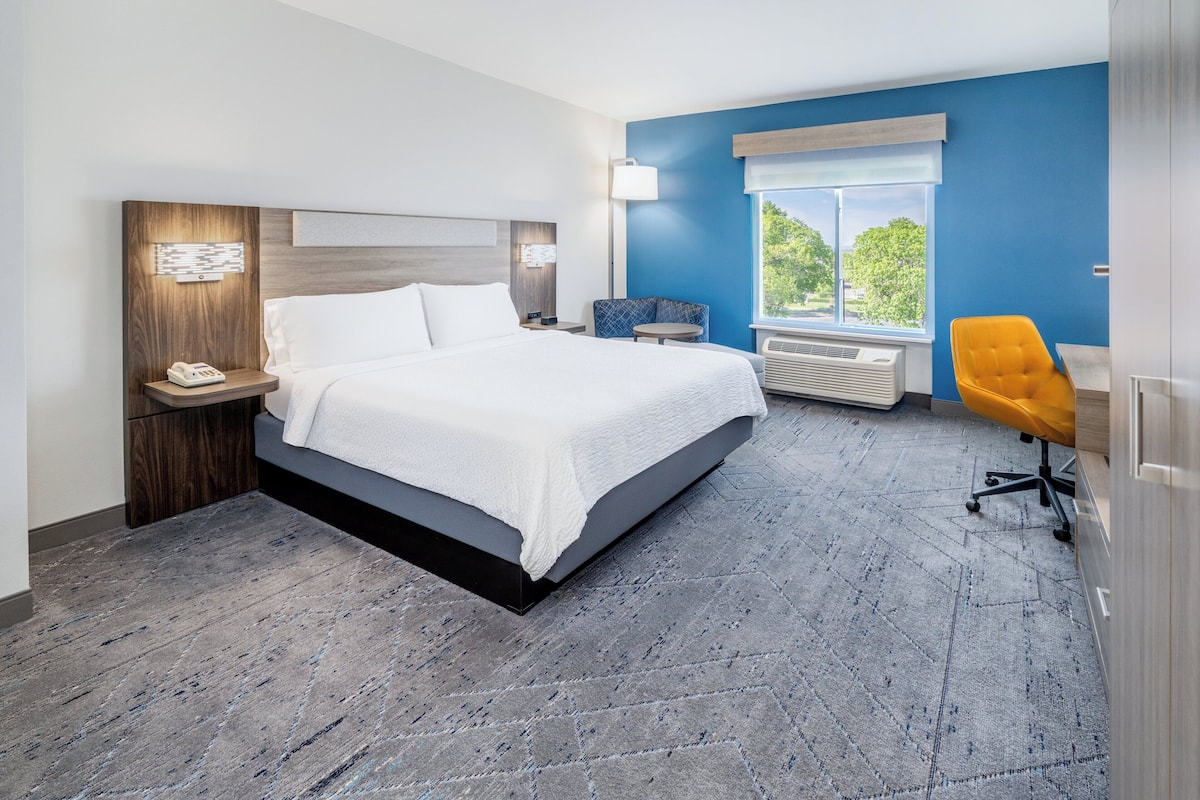 A modern hotel room with a large bed, a desk, a chair, and a window with a view of greenery.