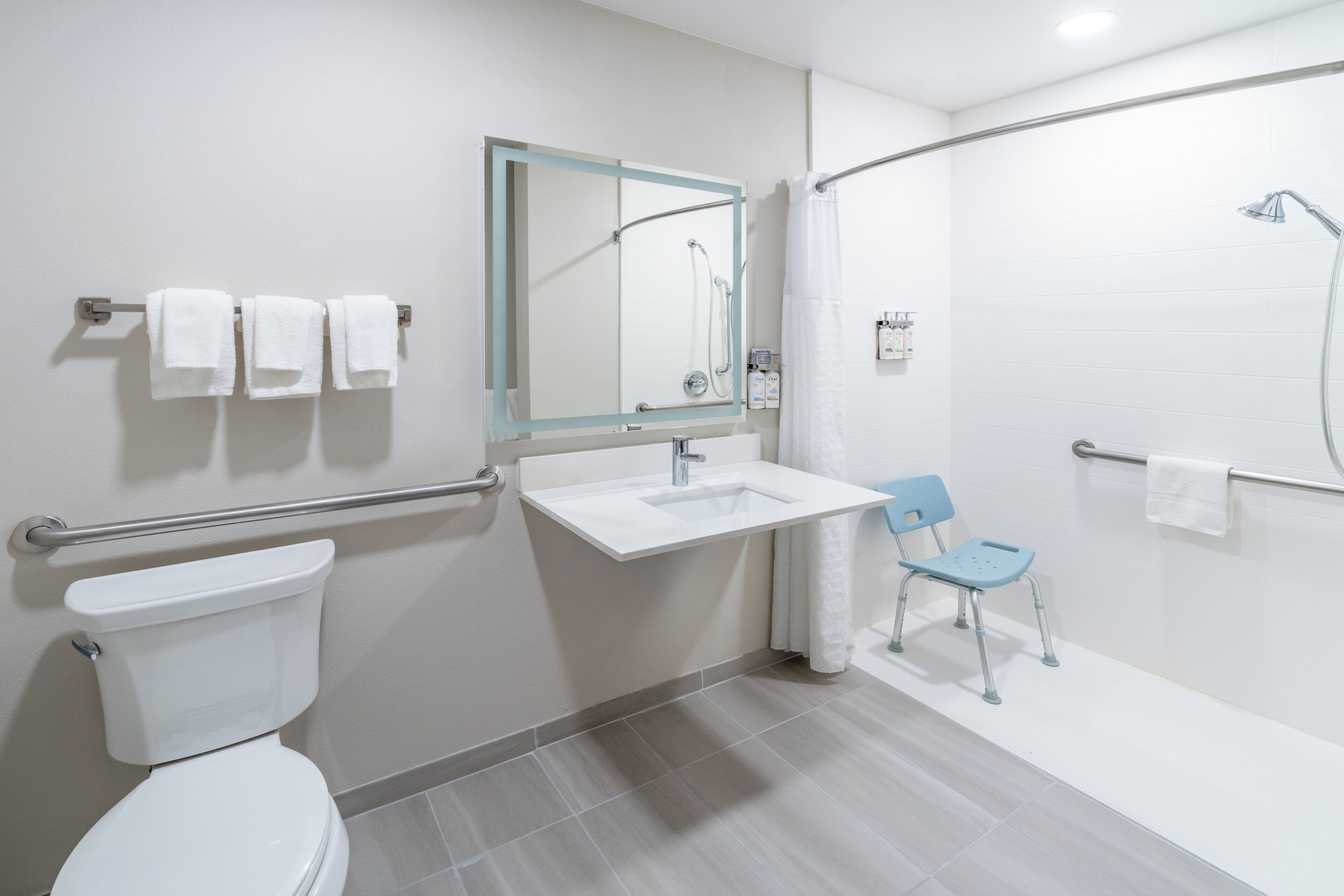 A modern bathroom with a toilet, sink, and shower. There is a chair and a handrail.