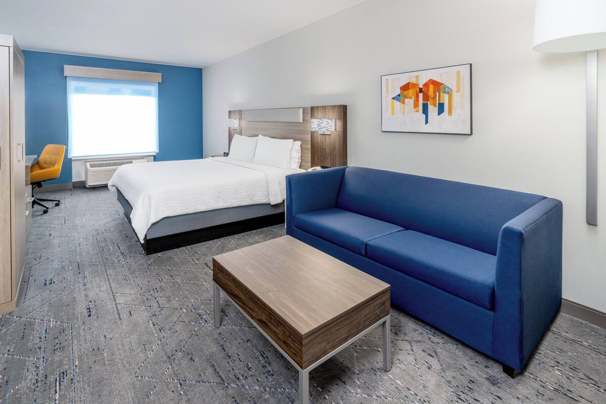 A hotel room with a bed, a blue sofa, a wooden coffee table, a desk with a chair, and a window with blinds.