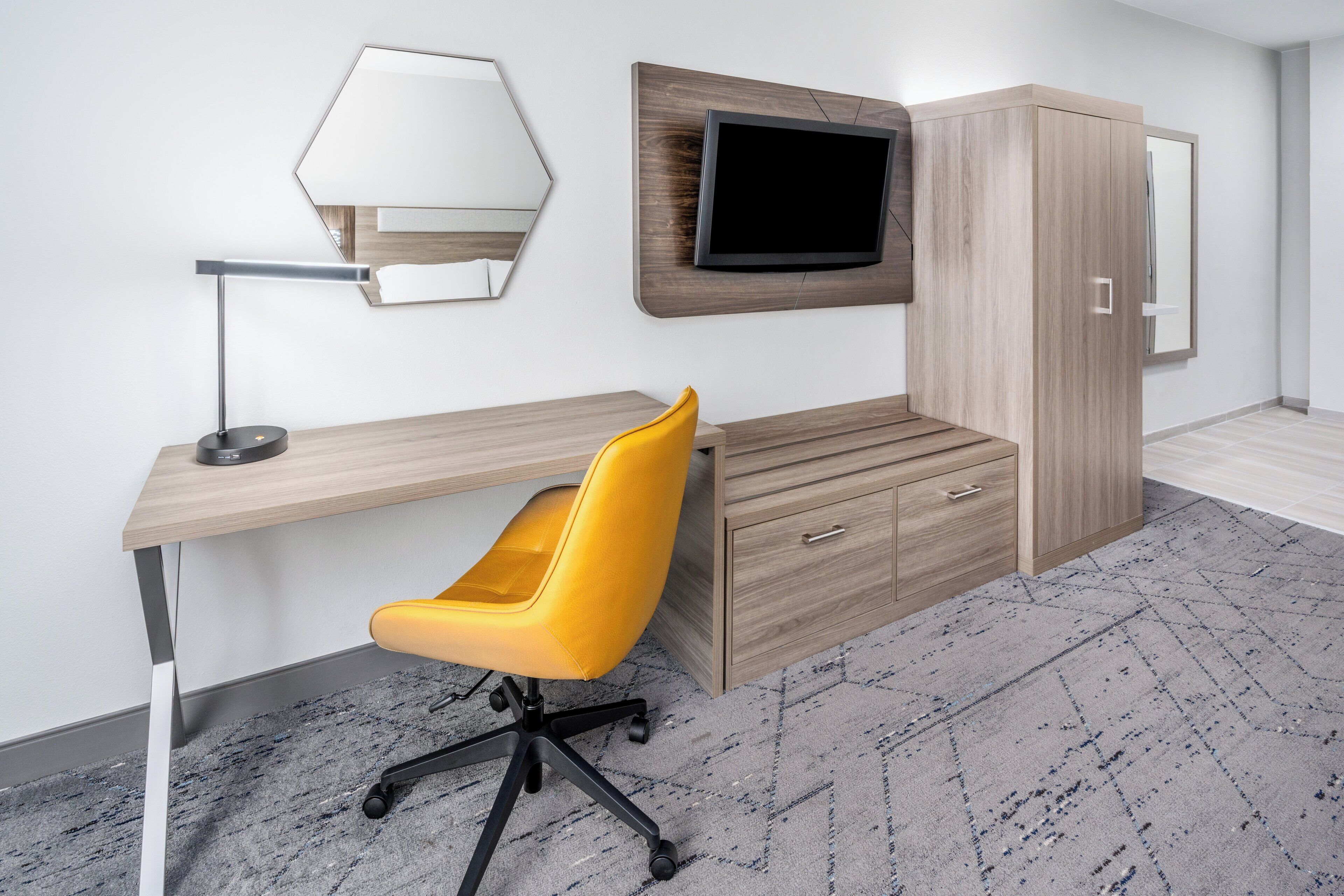 A modern hotel room with a wooden desk, a yellow chair, a flat-screen TV mounted on the wall, and a wardrobe.