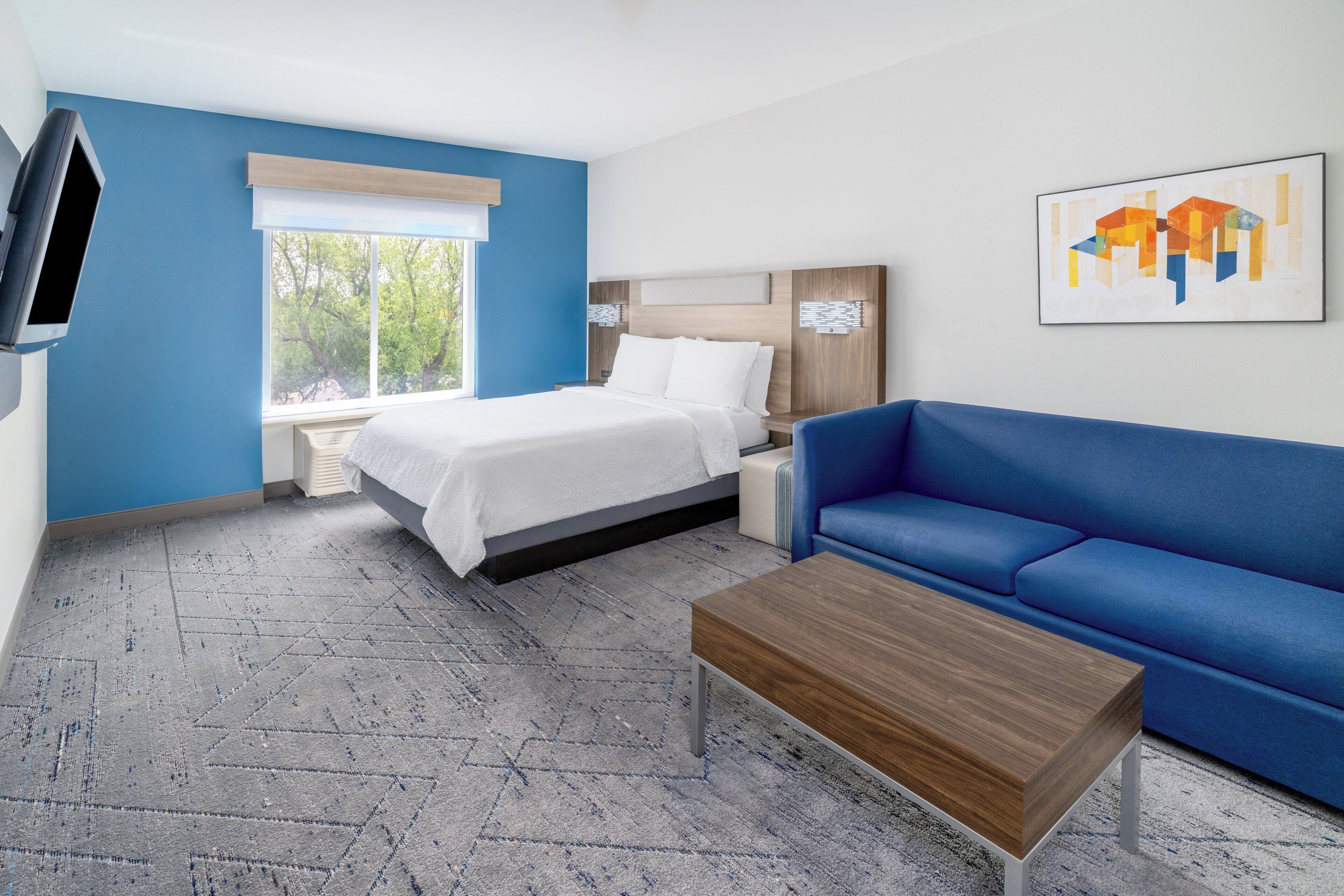 Een moderne hotelkamer met een bed, een blauwe bank, een houten salontafel en een aan de muur bevestigde televisie.