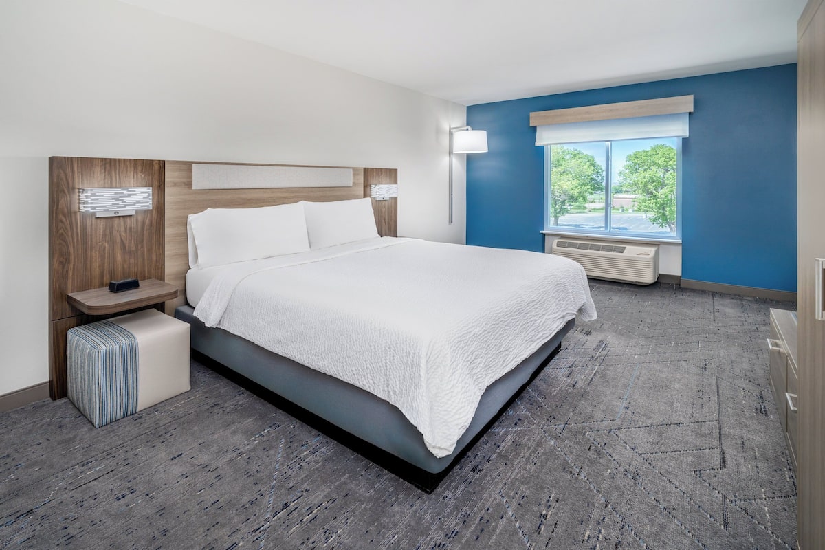 A modern hotel room with a large bed, a bedside table with a lamp, a window with a view of trees, and a built-in air conditioning unit.