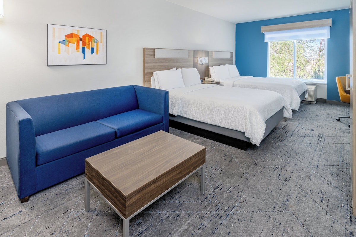 A hotel room with a blue sofa, a wooden coffee table, two beds, a nightstand, and a window with a view of trees.