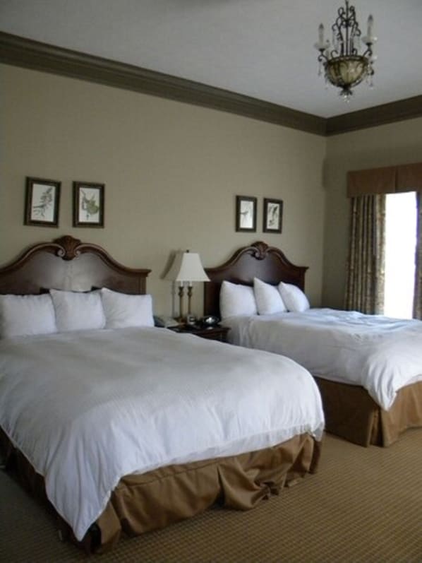 A hotel room with two beds, a chandelier, framed pictures on the wall, and a window with curtains.