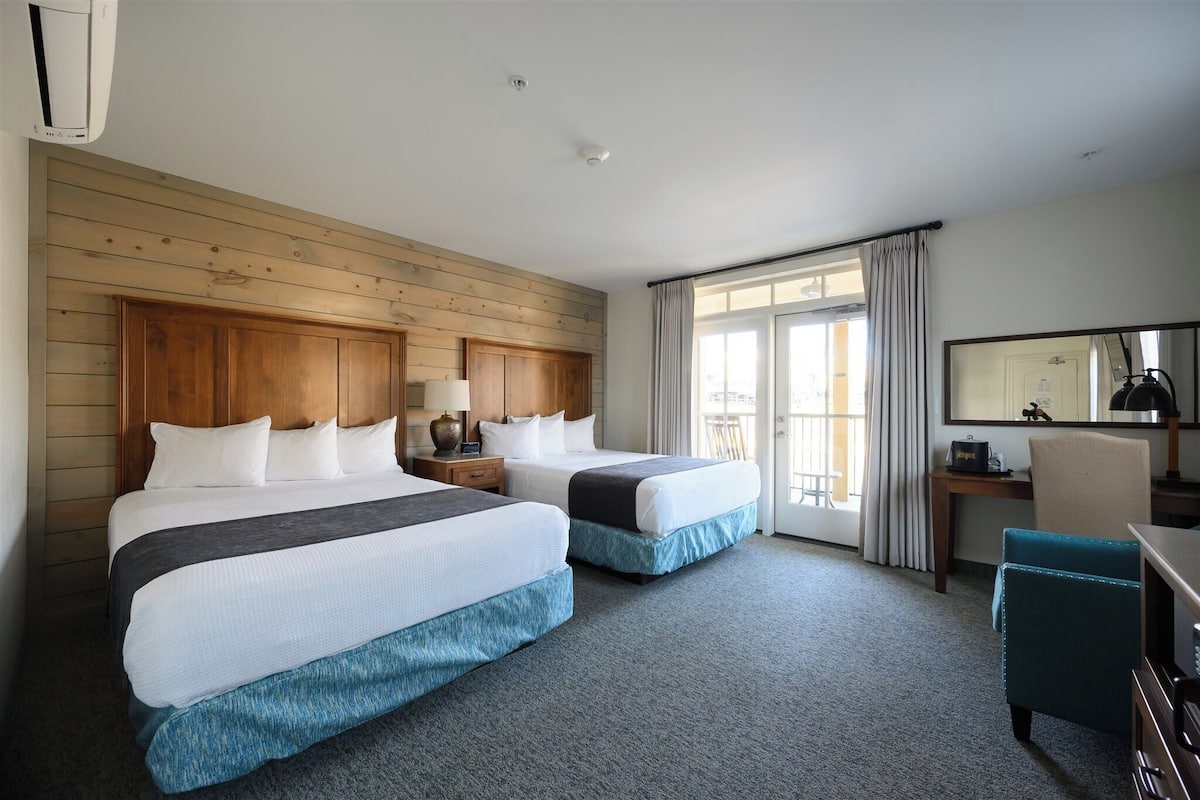 Quarto de hotel com duas camas, painéis de madeira nas paredes, uma escrivaninha com cadeira e uma janela grande com cortinas.