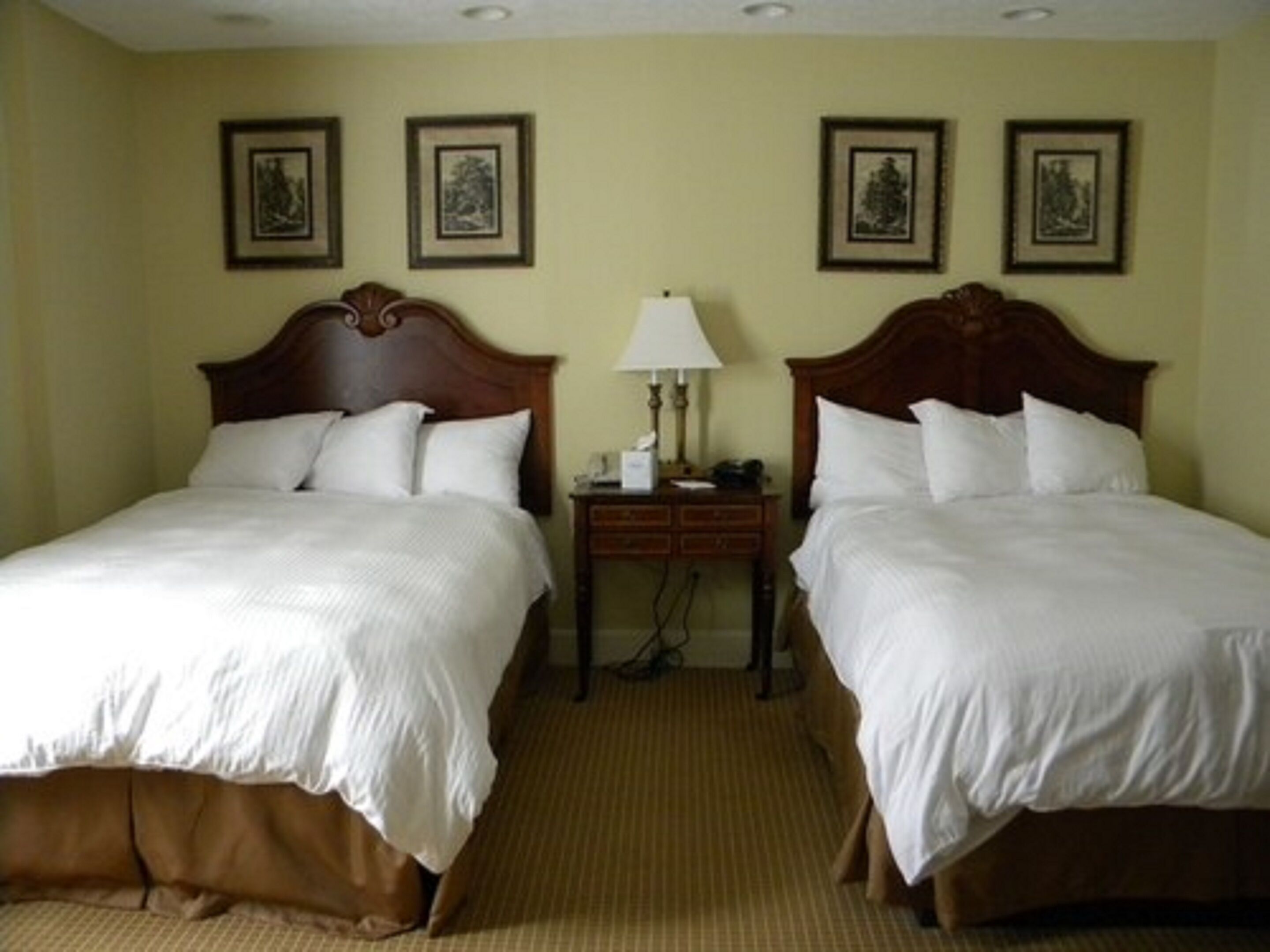 A hotel room with two beds, each with a wooden headboard, a bedside table with a lamp, and framed pictures on the wall.