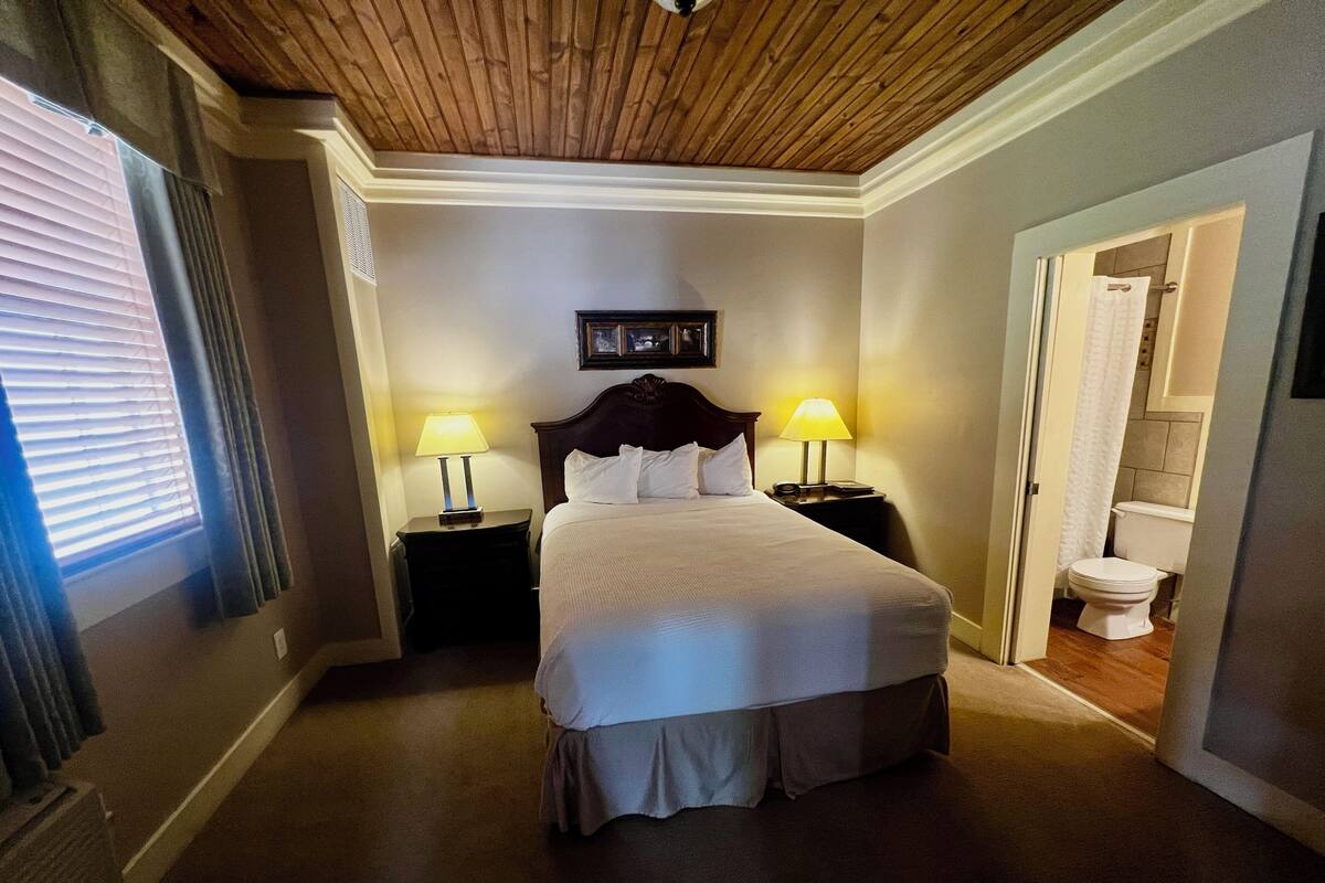 Quarto de hotel com teto de madeira, cama com roupa de cama branca, duas luminárias de cabeceira e vista para o banheiro.
