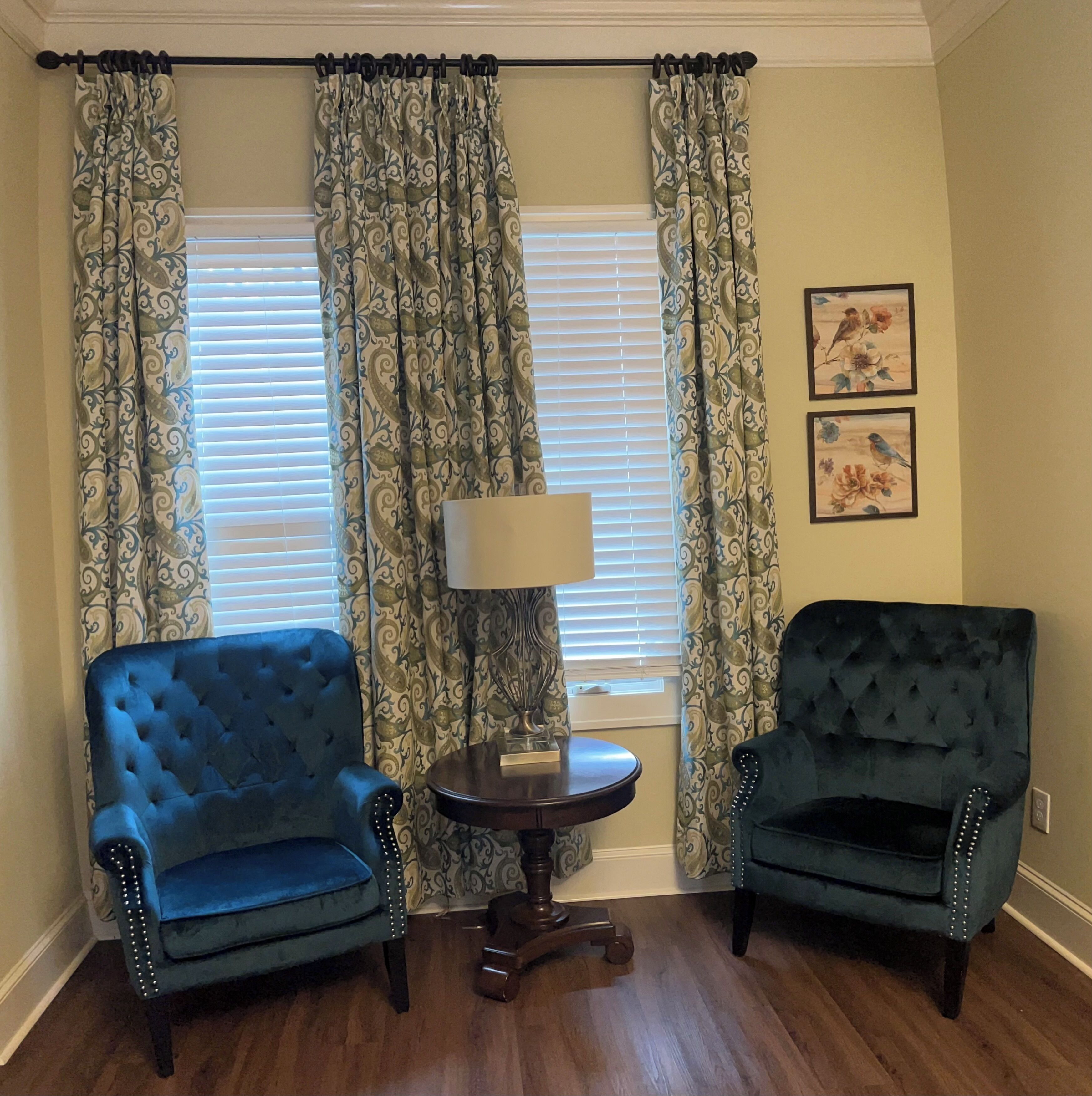 Een kamer met twee fauteuils, een bijzettafeltje met een lamp en een raam met zonwering en decoratieve gordijnen.