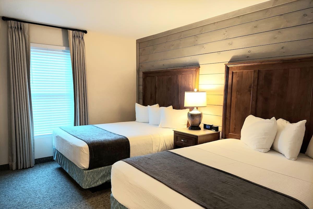Quarto de hotel com duas camas, uma mesa de cabeceira com abajur e uma janela com cortinas.