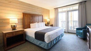 Une chambre d’hôtel avec une tête de lit en bois, un grand lit avec du linge de lit blanc, deux tables de chevet avec des lampes, un fauteuil vert et une vue sur un balcon extérieur, accessible par une porte vitrée.