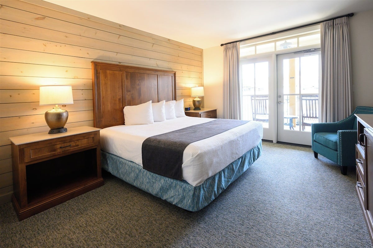 Quarto de hotel com cabeceira de madeira, uma cama grande com lençóis brancos, duas mesas de cabeceira com abajures, uma poltrona verde e vista para uma varanda através de uma porta de vidro.