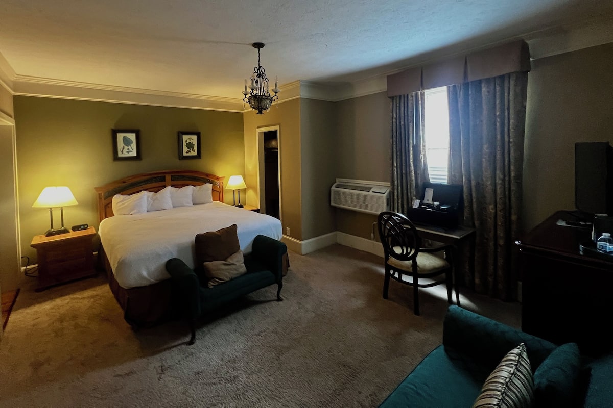 Quarto de hotel com cama, abajures, escrivaninha com cadeira, televisão e janela com cortinas.