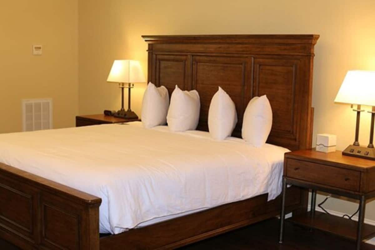 Cama bem arrumada com lençóis brancos e cabeceira de madeira, com duas mesas de cabeceira e luminárias de cada lado.