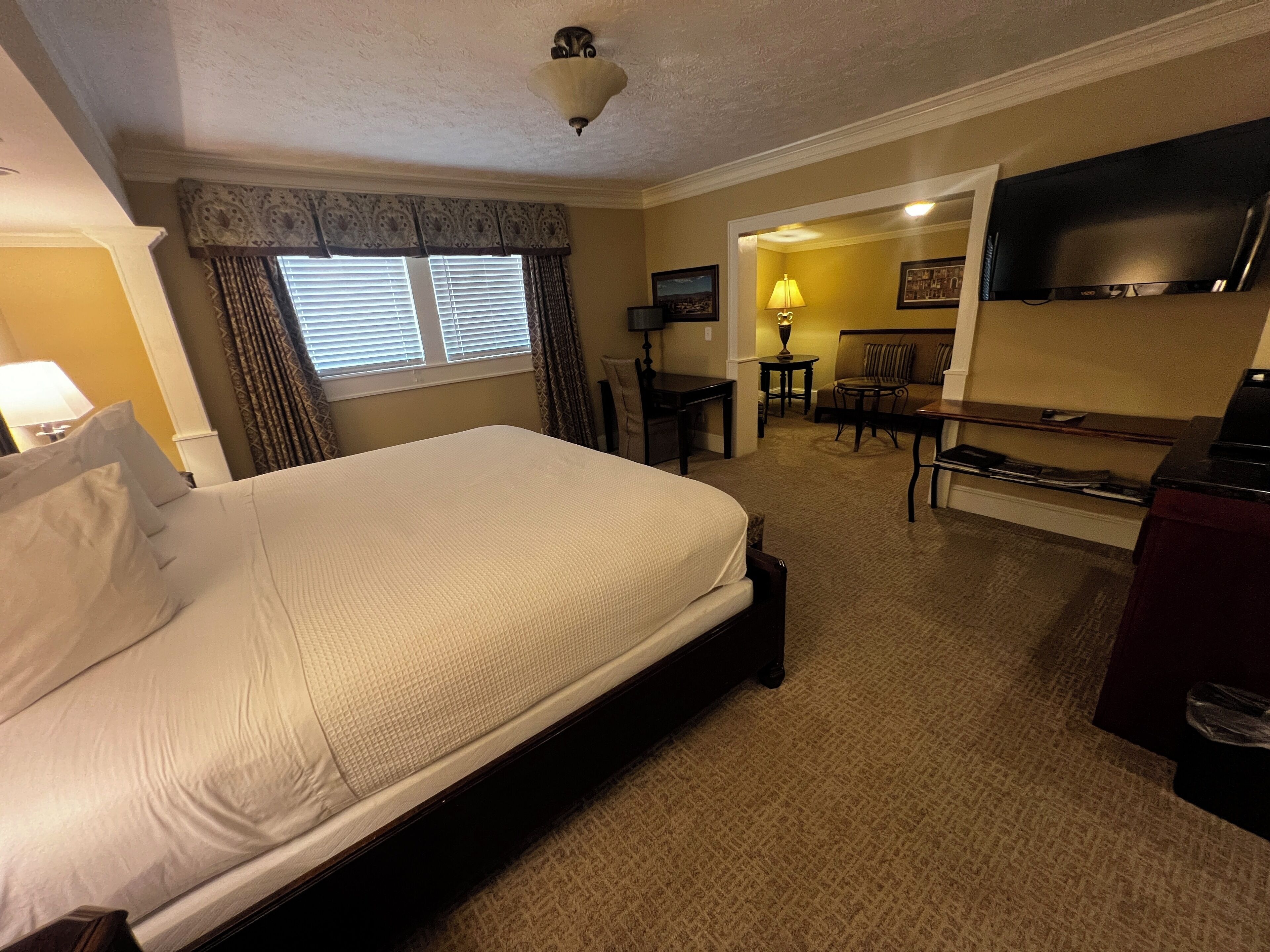 A hotel room with a large bed, a desk, a chair, a TV, and a lamp.