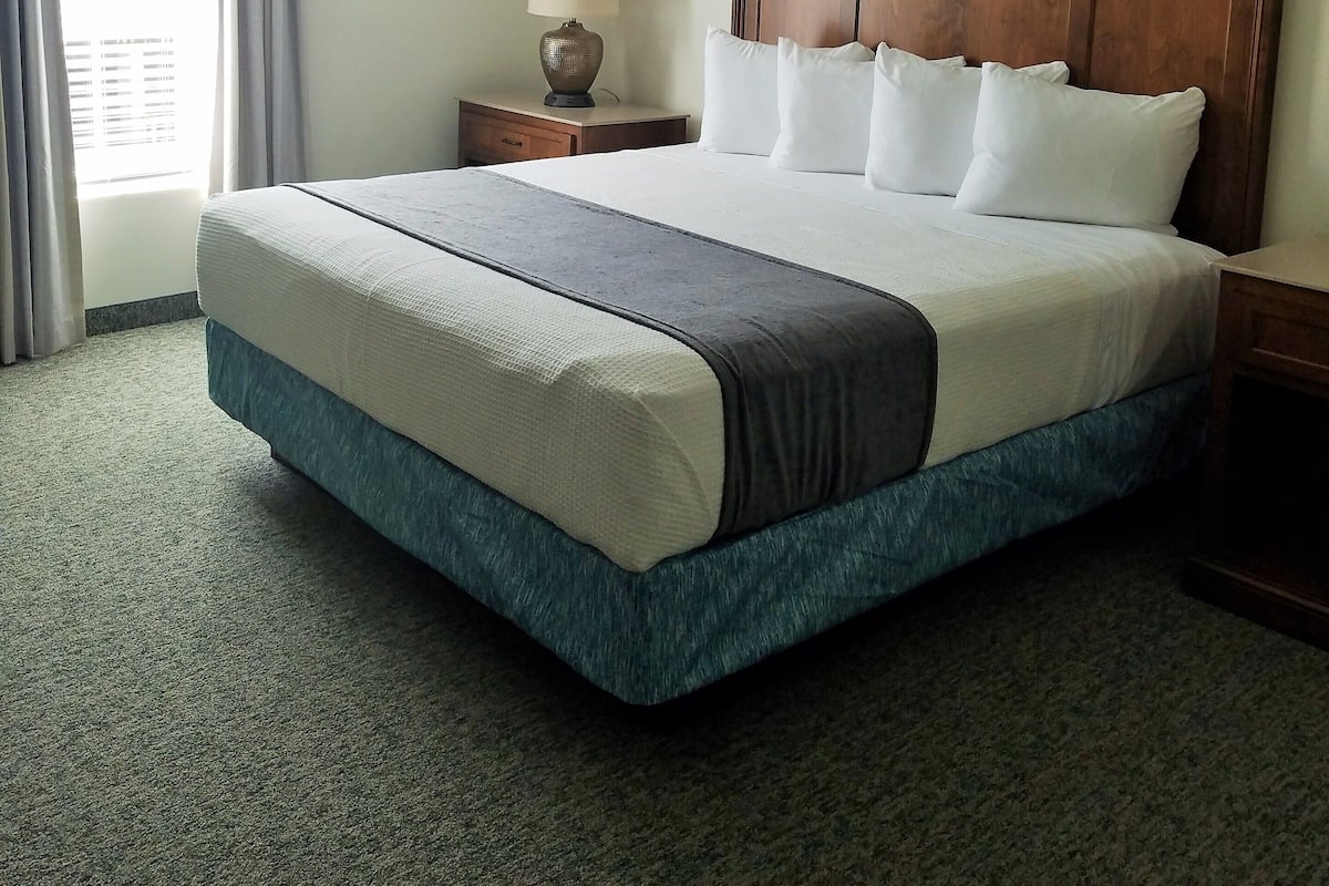 Quarto de hotel com uma cama grande, cabeceira de madeira, mesa de cabeceira com luminária e janela com cortinas.
