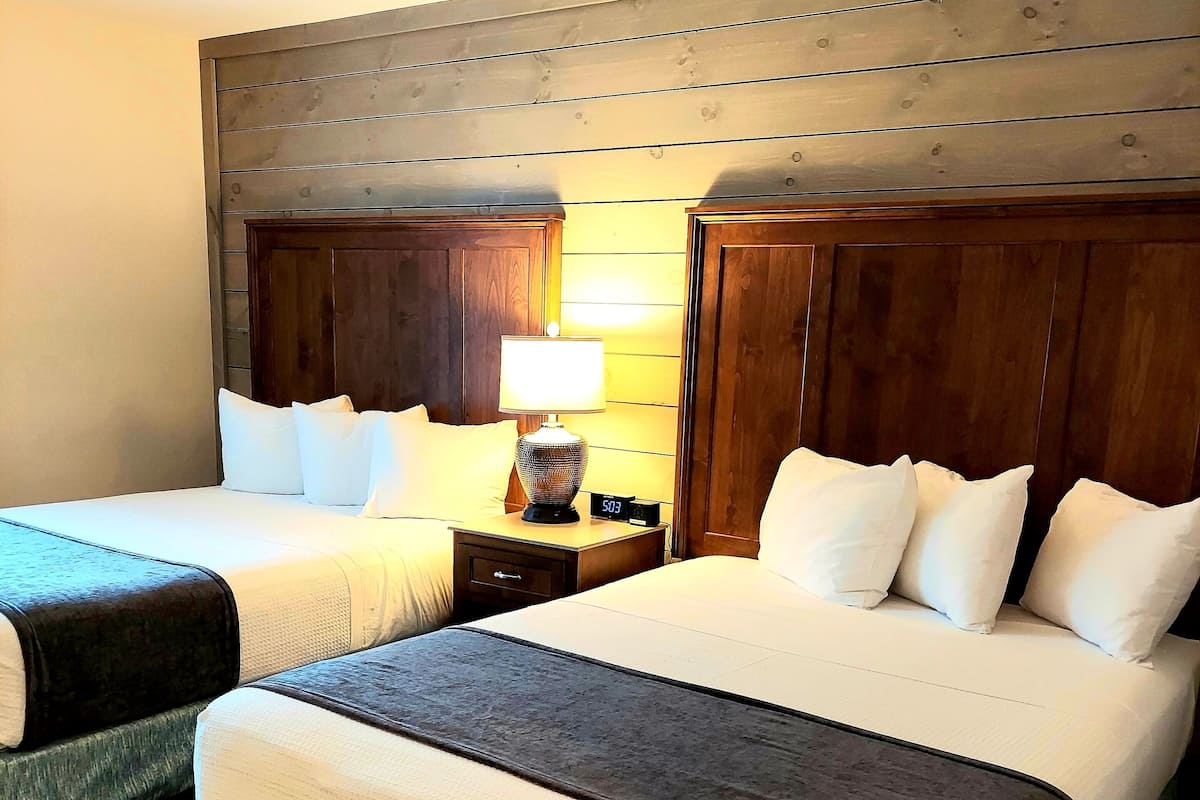 Quarto de hotel com duas camas, cabeceiras de madeira, mesa de cabeceira com luminária e relógio digital.