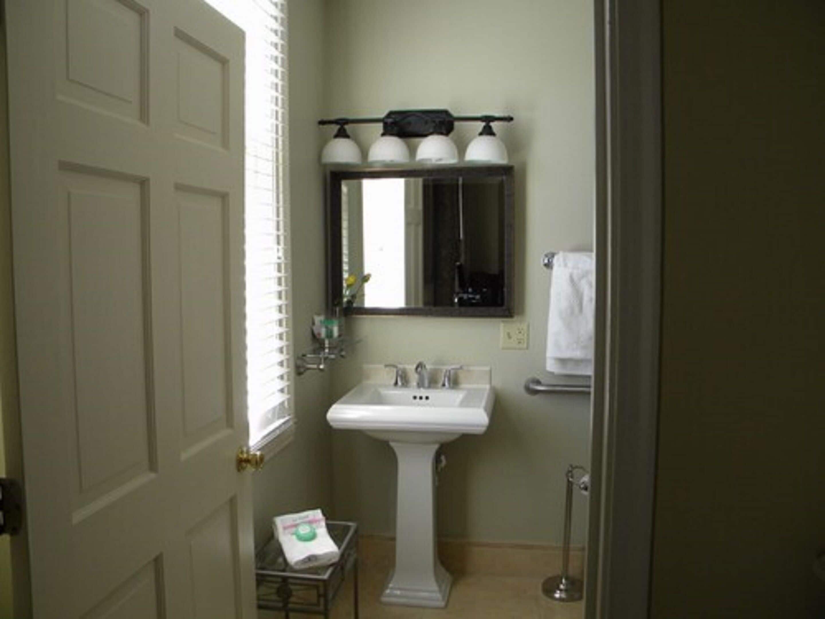 Bagno con lavabo su piedistallo, uno specchio sopra, portasciuga e un piccolo tavolo con una bottiglia.