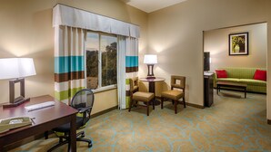 Une chambre d’hôtel équipée d’un bureau, de deux chaises, d’une table ronde avec une lampe, d’un canapé vert avec des coussins rouges et d’un tableau encadré au mur.