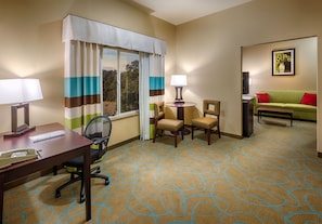 A hotel room with a desk, two chairs, a round table with a lamp, a green sofa with red cushions, and a framed picture on the wall.