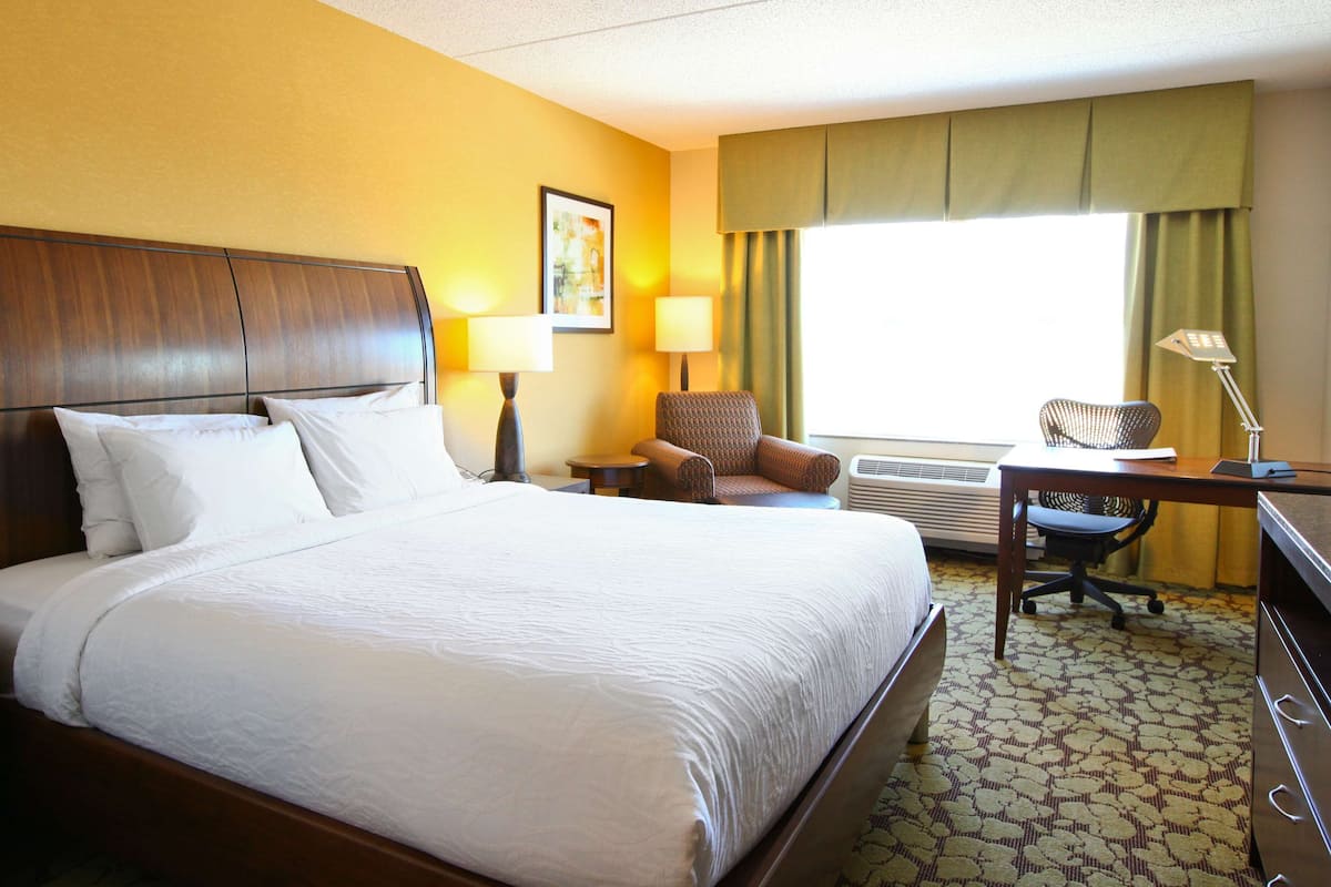 A hotel room with a large bed, a desk with a chair, an air conditioner, and a window with curtains.