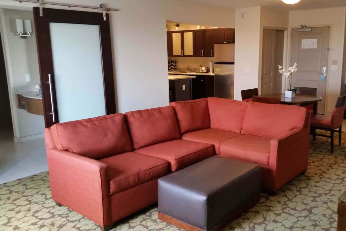 A living room with a sectional sofa, a small ottoman, and a kitchen in the background.