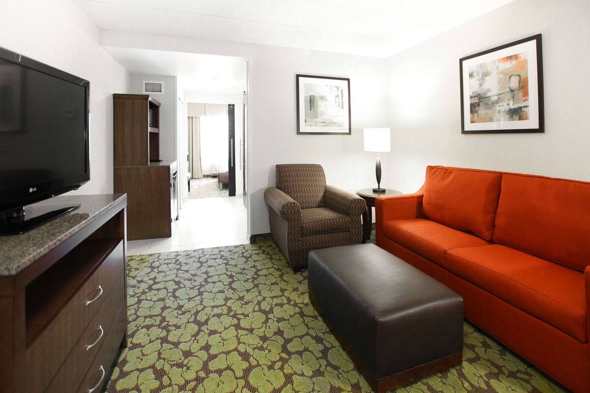 A hotel room with a television, a sofa, a chair, a small table with a lamp, and artwork on the walls.