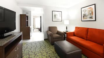 A hotel room with a television, a sofa, a chair, a small table with a lamp, and artwork on the walls.