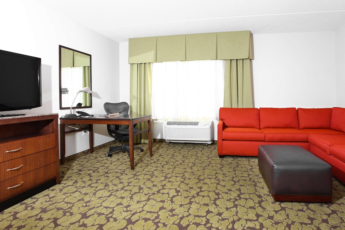 A hotel room with a television, desk, chair, red sofa, and an ottoman.