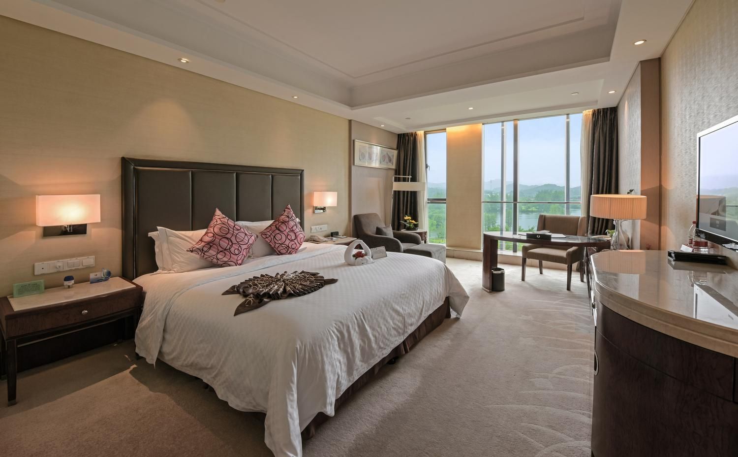 A hotel room with a large bed, a desk, a chair, a TV, and a balcony with a view.