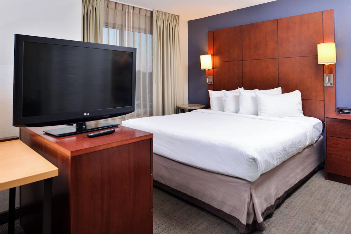 A hotel room with a large flat-screen TV, a wooden desk, a bed with white linens, and two wall-mounted lamps.