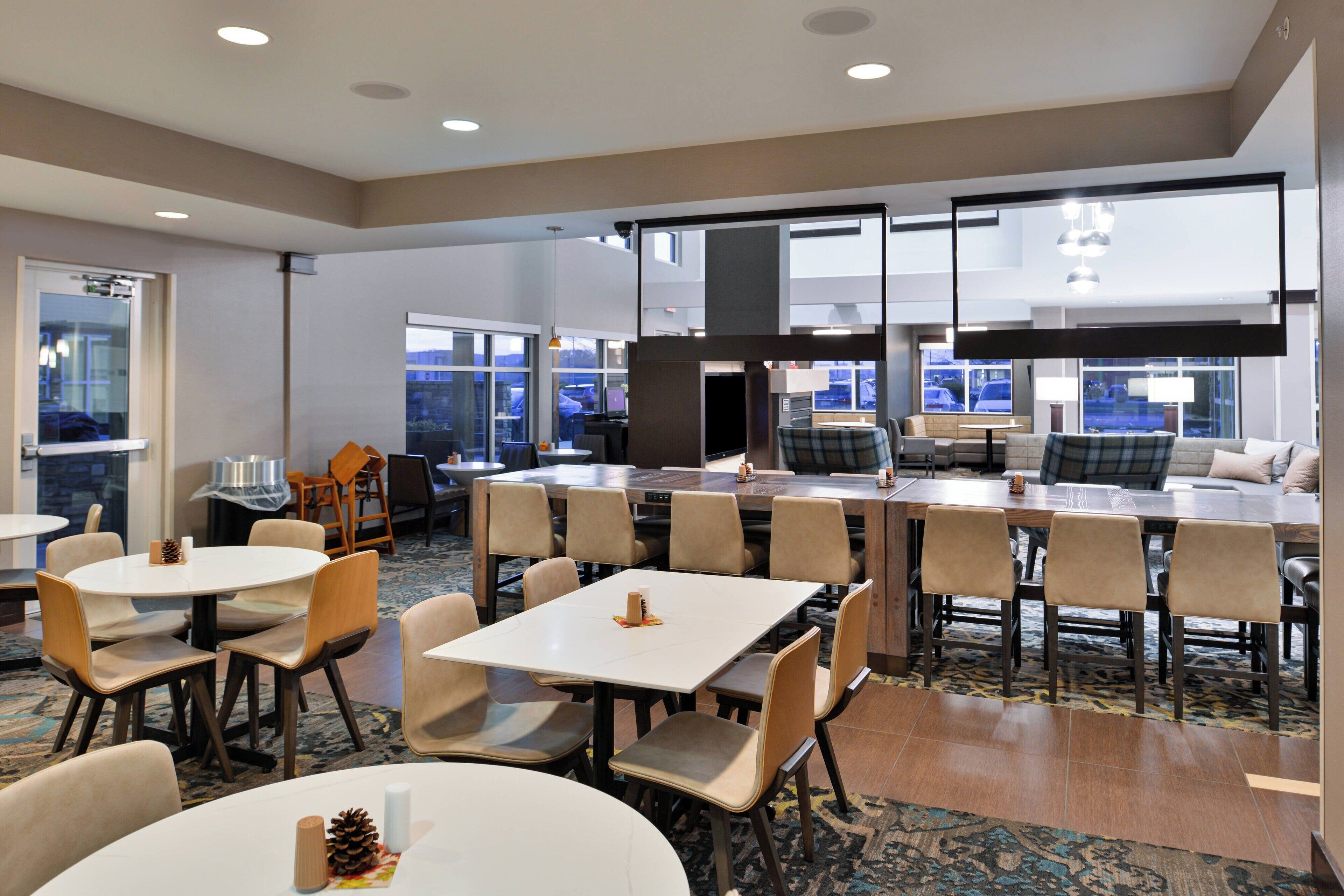 Un hall d’hôtel moderne doté de plusieurs espaces de détente, notamment des tables rondes et carrées avec des chaises, un canapé et une cheminée.