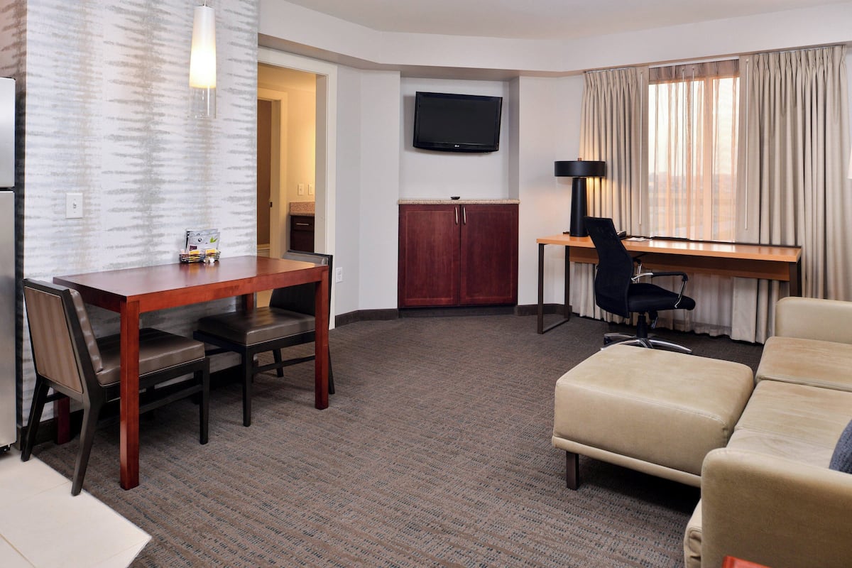 A hotel room with a wooden dining table, two chairs, a sofa, a desk with a chair, a flat-screen TV, and a large window with curtains.