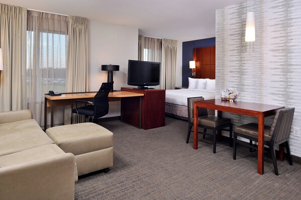 A hotel room with a bed, a desk with a television, a dining table with chairs, and a sofa.