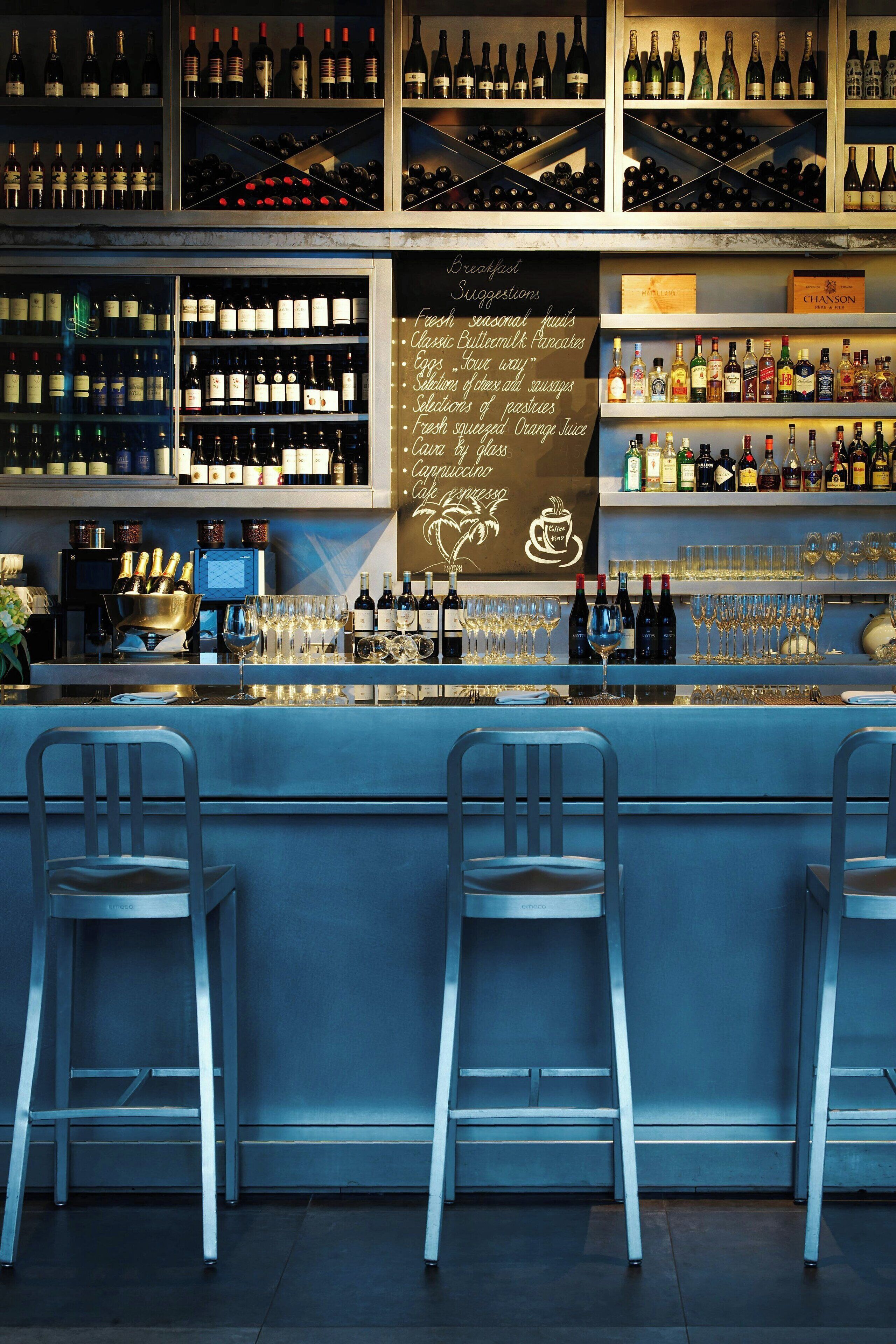 A bar area with multiple shelves of wine bottles, a chalkboard with breakfast suggestions, and bar stools.