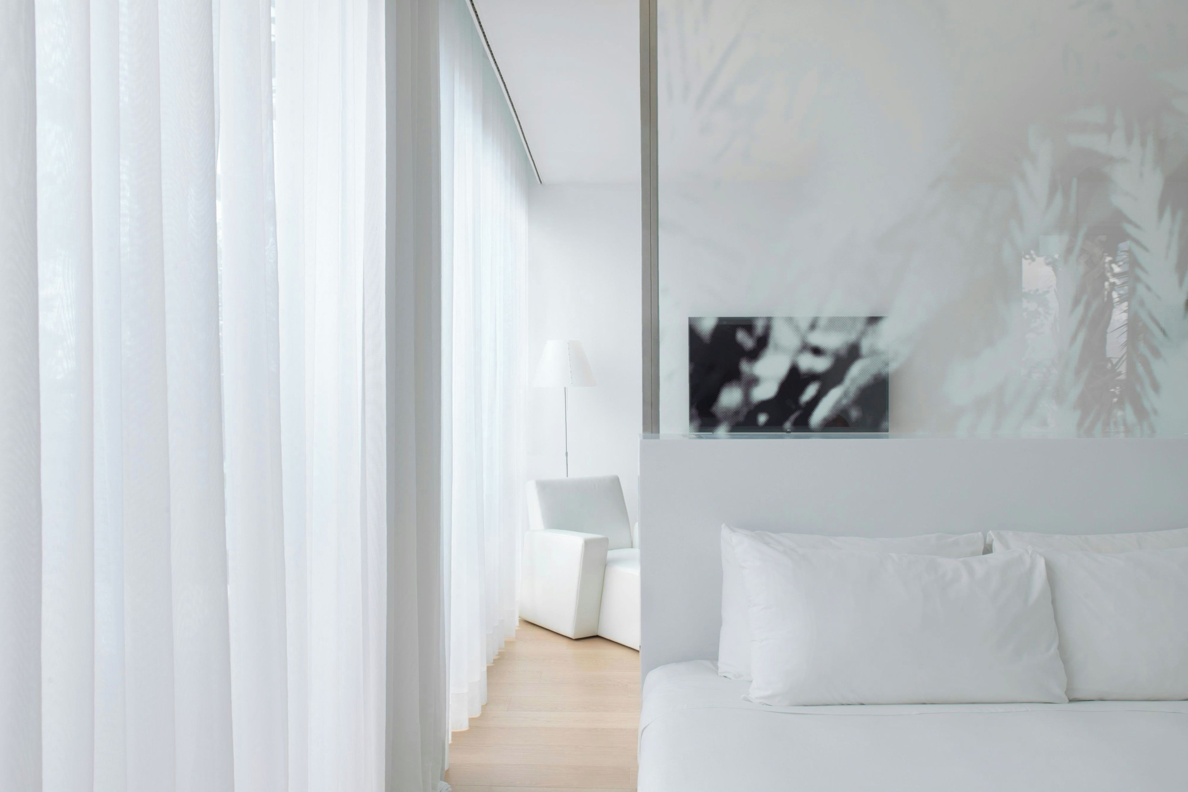A modern hotel room with a bed, white curtains, and a wall-mounted artwork.